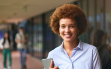 mulher negra com cabelo black power ruivo, camisa de botão azul segurando um tablet, em um ambiente aberto de escola.