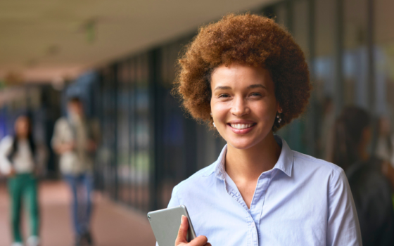 mulher negra com cabelo black power ruivo, camisa de botão azul segurando um tablet, em um ambiente aberto de escola.