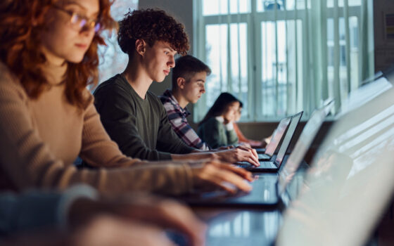 Alunos do ensino médio sentados em uma fileira, cada um com seu notebook.