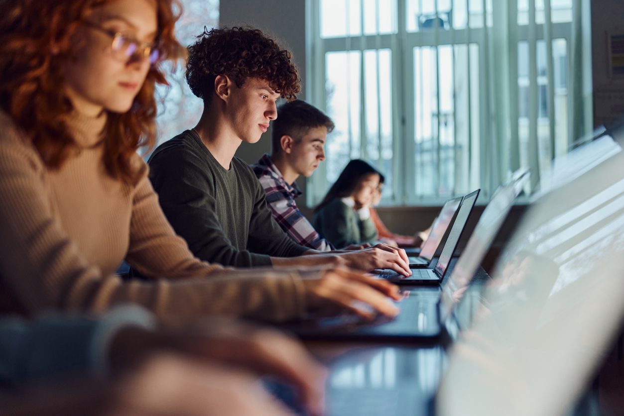 Alunos do ensino médio sentados em uma fileira, cada um com seu notebook.